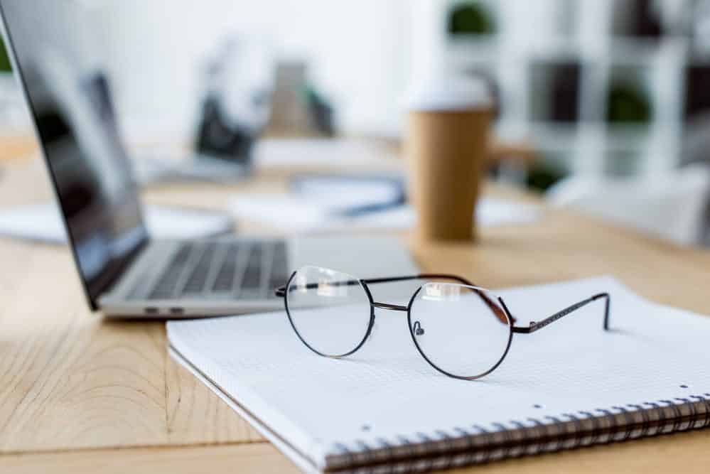 Laptop on desk with a notebook and pair of glasses.